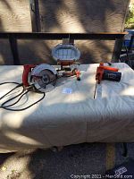 Overview of all three tools laid out on a white cloth table surface including chainsaw, Skilsaw, and work lamp