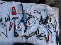 Overhead image of approximately 50 assorted hand tools spread out on white surface, showing staple guns, pliers, wrenches, hammer, tape, and cutters.