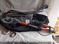 Tacklife disc sander with orange and black handle and flexible dust collection hose, sitting on top of a black carrying bag. Also shown are rolls of tape next to the sander setup.