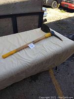 Full view of the Garant double ended pickaxe with wooden handle laid on a beige surface outdoors.
