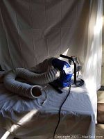 Photo showing blue dust collection blower with white flexible hose on a white cloth backdrop. Black metal handle frame and power cord visible.