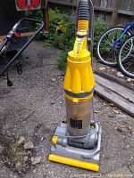 Front angle view of the Dyson vacuum cleaner standing on outdoor gravel with the dust canister clearly visible. Yellow and gray colors with cord extended on the ground.