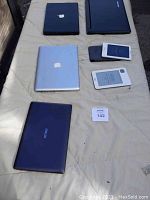 Closed black Apple MacBook and closed Lenovo laptop at top, closed silver Apple MacBook, black and white e-readers in protective cases, closed Asus laptop at bottom