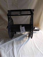 Black metal garden hose holder laying on white cloth background, side perspective showing decorative cutout side panels and mounting bracket on top