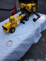Photo showing two metal Tonka sandbox trucks, a motor grader and an excavator, on a white sheet outdoors with visible wear and detail