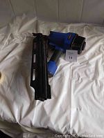 Blue and black Campbell Hausfeld pneumatic nailer placed on white surface with a lot number card beside it.