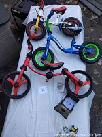 Three children's balance bikes laid on a white sheet outdoors along with some unrelated items. Shows red, blue, and white balance bikes.
