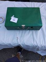 Closed green metal box containing the camping stove, with latch and handle visible.