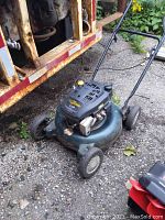 Front-angle view of Yardworks lawn mower showing the black engine cover, green deck, and front wheels on rough ground.