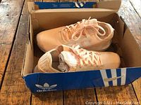 Pair of light peach Adidas running shoes in original blue Adidas box on wood floor.