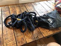 Expandable black garden hose coiled loosely on a wooden table with brass connectors visible. Next to it is a black plastic storage bag with original barcode attached.