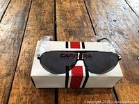 Sunglasses with dark tinted lenses and black frames resting on a white Carrera branded box with red and black stripes on a wooden surface.