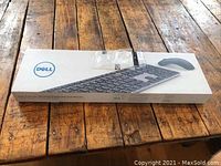 Boxed Dell Premium Wireless Keyboard and Mouse set shown on a wooden surface with visible Dell branding and product images on the packaging.