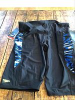 Front view of black Speedo competitive swim shorts with blue side panels and visible Speedo logo, displayed flat on a wooden surface.