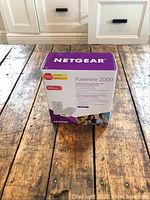 Box of Netgear Powerline 2000 adapters on wooden floor with white cabinetry in background, showing purple and white packaging with product usage information.