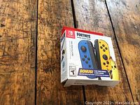 Sealed box showing front of Nintendo Switch Fortnite Edition Joy-Con controller set, blue left controller and yellow right controller, shown on wooden surface.