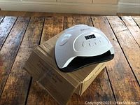 White SUN X7 80W LED nail polish gel dryer lamp on a brown cardboard box labeled 'Professional Gel Polish LED Nail Dryer Lamp' on a wooden surface.