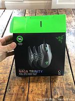 Razer Naga Trinity gaming mouse box held in hand showing front with image of mouse and branding details on wooden surface