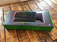 Photo of the sealed Razer Huntsman Elite gaming keyboard box on wooden floor, showing front product image with RGB lighting on keys.