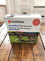 Front view of the unopened Gardena ZoomMaxX Sled Sprinkler box on a wooden surface showing product details and branding.