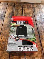 Photo of boxed Weber premium grill cover on a wooden floor, showing front package graphics with Weber logo and image of cover on grill.