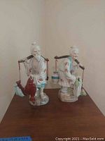 Front view of both Asian porcelain statues, male on left holding fish, female on right carrying objects
