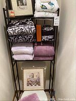 Full view of black metal bathroom rack holding towels, framed art, candle, and box.