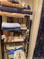 Towels stacked on the black metal over-the-toilet rack with shelves holding air fresheners, wipes, and other bathroom items, with Dick's Vaporizer box on lowest shelf