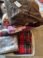 Image showing a stack of assorted blankets including a brown faux fur blanket, quilted patterned blankets, and a red and green plaid blanket.