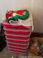 Stack of eight red plastic holiday storage bins with lids. Tree skirt partially placed on top.