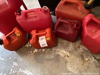 Seven red and orange plastic gas cans of various sizes and shapes sitting on a concrete floor.