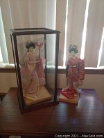 Wide view showing both Geisha dolls, one inside a glass display case and the other on wooden stand outside case, on wooden table.