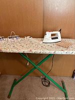 Photo of the ironing board with floral cover, green legs, white iron on top, two white holders and wooden clothespins