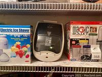 Photo showing Electric Ice Shaver box, George Foreman Lean Mean Fat Grilling Machine, and Magic Bullet Express set on shelf.