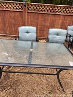 Outdoor rectangular table with textured glass top and black metal frame, showing two of the cushioned metal chairs behind it.
