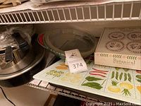 Stainless steel stock pot with lid, clear glass roasting pan, a box of Botanical by Home Essentials plates, kitchen linen towel with vegetable designs, stored on a shelf.