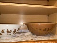 Two vintage Pyrex mixing bowls side by side on a shelf. Left bowl is white with brown rooster/weather vane design, right bowl is brown with similar pattern.