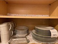 Cabinet shelf containing white dishes with gold trim including stacked dinner plates, stacked side plates, stacked bowls, stacked mugs, and a glass butter dish.