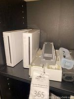 Two white Nintendo Wii consoles on a shelf with accessories including a sensor bar and remote covers.