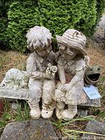 Statue of boy and girl sitting on a bench with a dog beside them and a frog garden ornament with a wide open mouth.