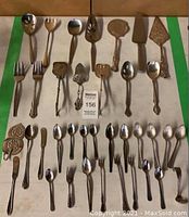 Full lot of 37 assorted utensils arranged in rows on a beige surface. Includes serving spoons, pie servers, forks, small spoons, and knives.