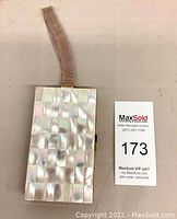 Closed rectangular clutch with mother of pearl tiled face in checkerboard pattern with attached wrist strap, placed next to auction label.