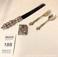 Four-piece lot includes two sterling silver spoons, silver serving cutter with ornate handle, and decorative silver matchbox. Items arranged on white flat surface with auction tag 188.