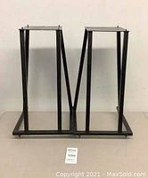 Front view of pair of black metal speaker stands with square top plates and visible wear, one missing a metal foot on base.