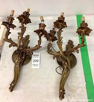Pair of brass wall sconces laid on a flat surface, showing the full front and detailed design with three arms each.
