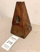 Front and side view of the antique Swiss wooden metronome showing its pyramid shape, metal emblem, and winding key with visible surface wear.
