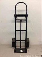 Front view of black metal 4 wheel convertible dolly in upright 2-wheel dolly mode, showing handlebar, base plate, and inflated rubber tires.