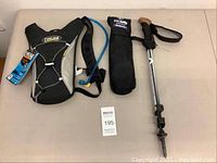 Three items laid out: Camelbak hydration pack with blue tube, black Pacsafe anti-theft cable in carrying case, and extendable Black Diamond walking stick with foam handle.