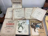 Overall view of several vintage sheet music books and loose sheets laid out on a table, showing different titles and styles in worn condition