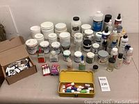 Overview of assorted used paint jars, bottles, and tubes on a table, including a yellow metal case with paint tubes and some cardboard boxes.